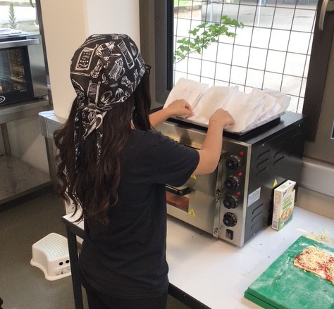 eine Schülerin steht vor dem Pizzaofen und legt die verpackten Pizzastücke oben auf den Ofen © Nora Leiner